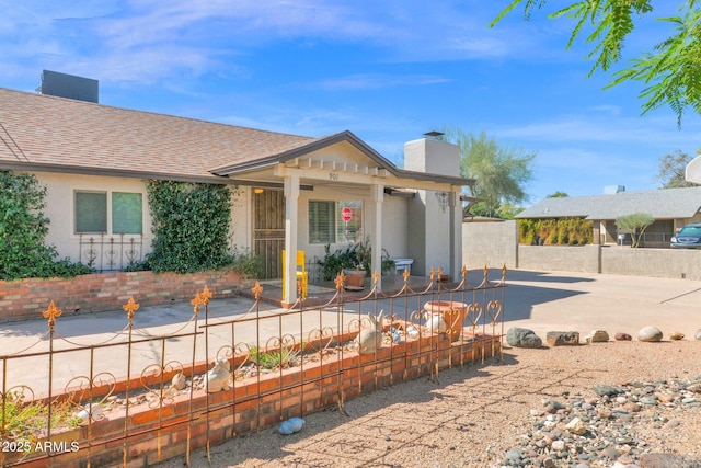 view of front facade with fence, roof with shingles, stucco siding, a chimney, and a patio area