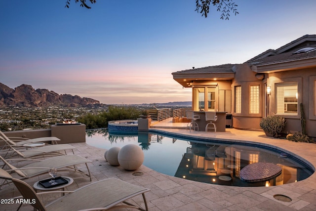 pool at dusk featuring an in ground hot tub, a mountain view, an outdoor bar, and a patio area