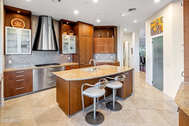 kitchen featuring a breakfast bar, stainless steel gas stovetop, an island with sink, sink, and wall chimney range hood