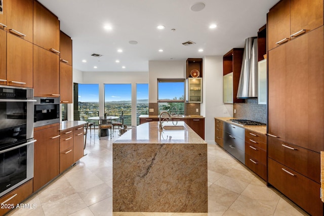 kitchen featuring wall chimney exhaust hood, light stone counters, a center island with sink, stainless steel appliances, and decorative backsplash