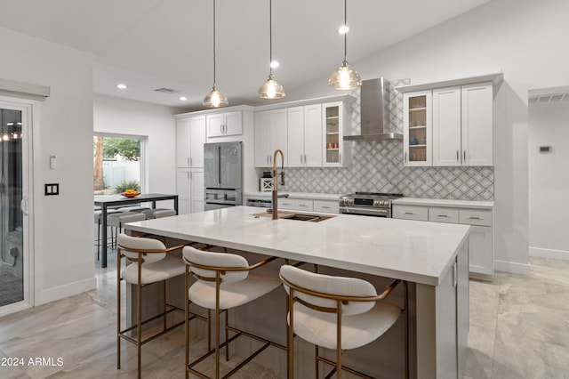 kitchen featuring sink, an island with sink, light stone countertops, wall chimney range hood, and appliances with stainless steel finishes