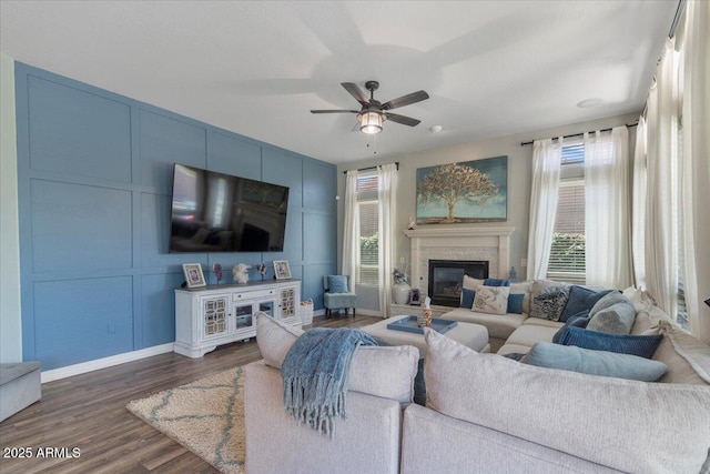 living area featuring a glass covered fireplace, a decorative wall, baseboards, ceiling fan, and dark wood-type flooring