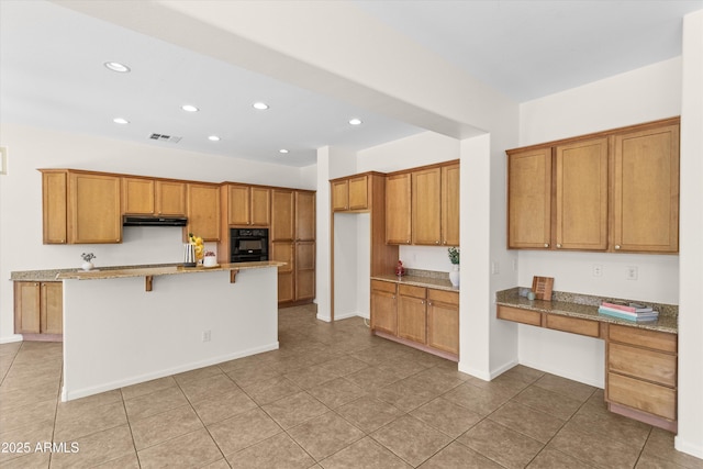 kitchen with under cabinet range hood, a kitchen breakfast bar, black oven, built in study area, and brown cabinetry