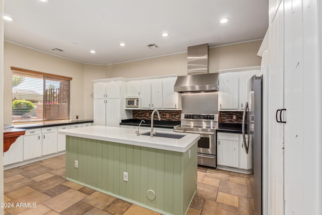 kitchen featuring backsplash, a kitchen island with sink, white cabinets, wall chimney range hood, and appliances with stainless steel finishes