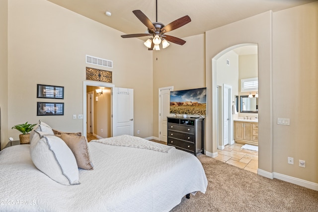 bedroom with ceiling fan, light colored carpet, ensuite bathroom, and a towering ceiling