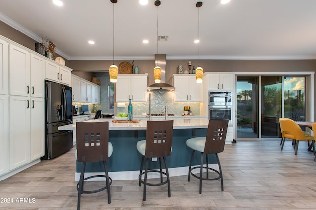 kitchen featuring wall chimney range hood, black fridge, an island with sink, hanging light fixtures, and white cabinets