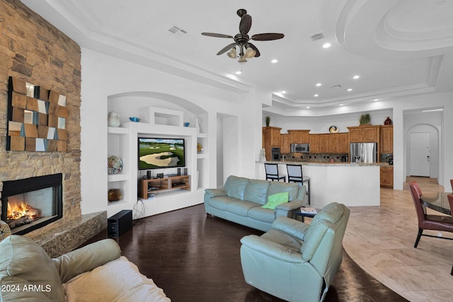 living room with ceiling fan, built in features, a tray ceiling, and a stone fireplace