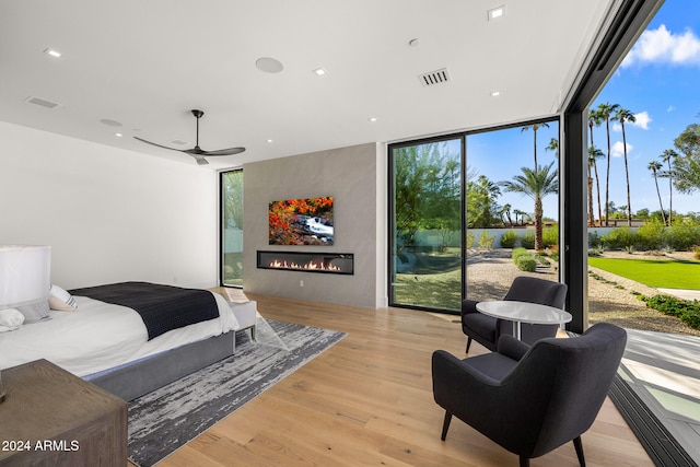 bedroom with a wall of windows, a large fireplace, ceiling fan, and light wood-type flooring