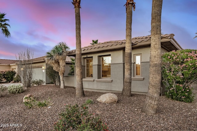 property exterior at dusk with a garage, a tiled roof, and stucco siding