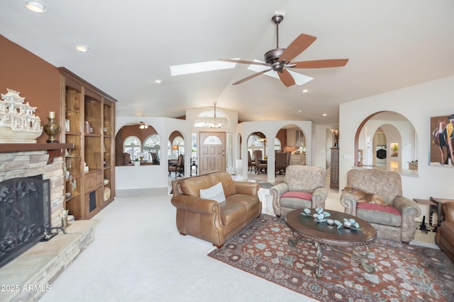 living area featuring arched walkways, lofted ceiling with skylight, light carpet, ceiling fan, and a stone fireplace