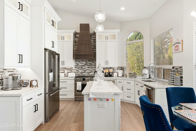 kitchen with appliances with stainless steel finishes, glass insert cabinets, a kitchen island, a sink, and wall chimney exhaust hood