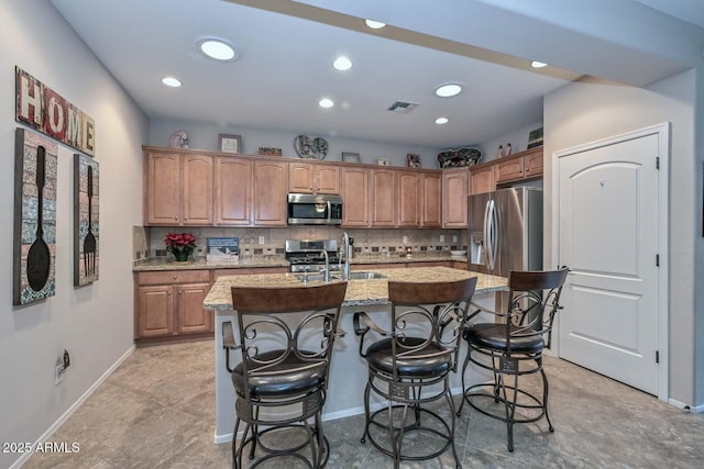 kitchen with a kitchen island with sink, sink, light stone counters, and appliances with stainless steel finishes