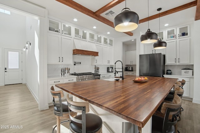 kitchen featuring appliances with stainless steel finishes, custom range hood, a kitchen island with sink, white cabinets, and beam ceiling