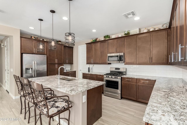 kitchen with light stone countertops, appliances with stainless steel finishes, sink, hanging light fixtures, and an island with sink