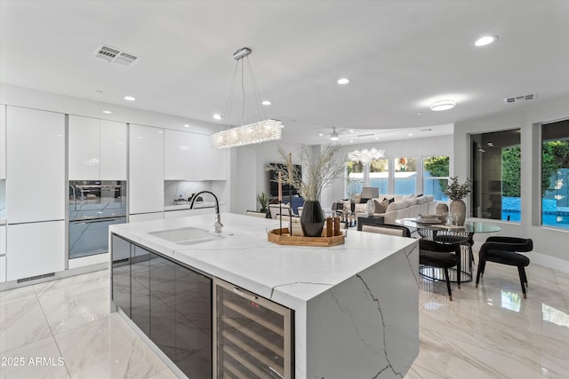 kitchen with decorative light fixtures, white cabinetry, wine cooler, sink, and a kitchen island with sink