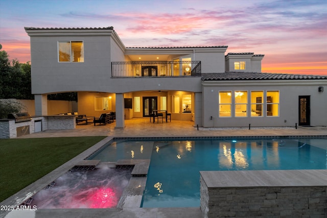 back house at dusk featuring ceiling fan, exterior kitchen, a balcony, a swimming pool with hot tub, and a patio