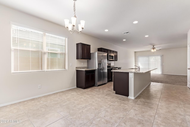 kitchen featuring light stone counters, decorative light fixtures, stainless steel appliances, an island with sink, and ceiling fan with notable chandelier