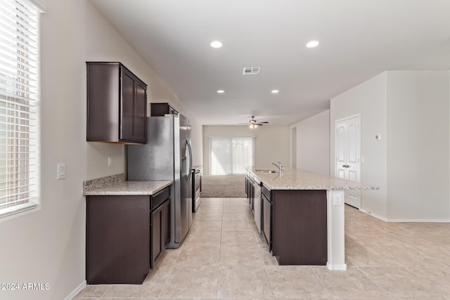 kitchen with light stone countertops, ceiling fan, a kitchen island with sink, and dark brown cabinetry