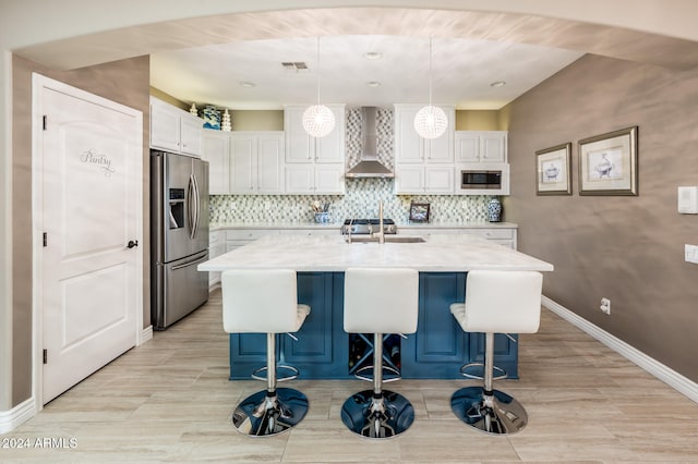 kitchen with wall chimney range hood, appliances with stainless steel finishes, an island with sink, white cabinetry, and decorative light fixtures