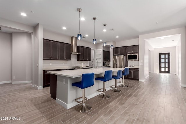 kitchen featuring wall chimney range hood, a kitchen breakfast bar, an island with sink, pendant lighting, and appliances with stainless steel finishes