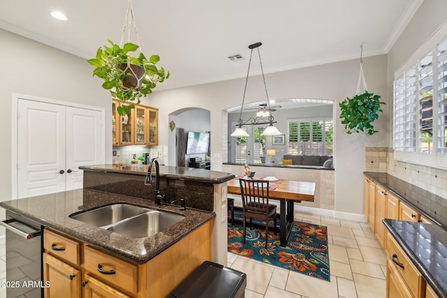 kitchen with tasteful backsplash, a sink, dark stone counters, arched walkways, and a kitchen island with sink