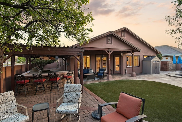 back of house at dusk featuring stucco siding, outdoor lounge area, a storage shed, an outdoor structure, and a patio