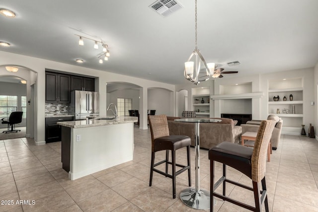 kitchen featuring arched walkways, a kitchen island with sink, dark cabinets, light stone countertops, and stainless steel fridge