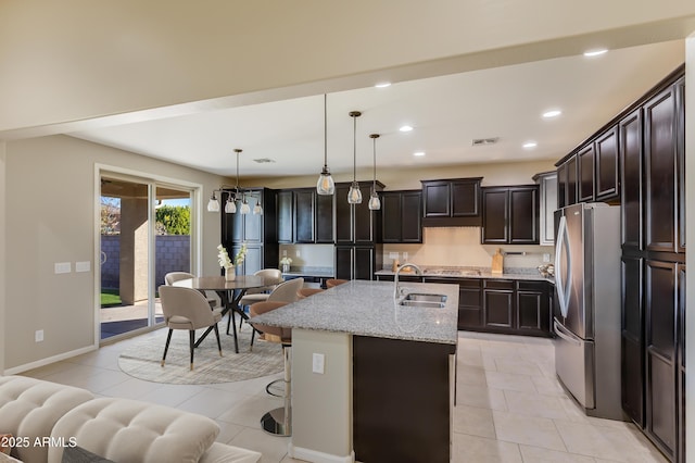 kitchen with sink, stainless steel fridge, hanging light fixtures, a kitchen island with sink, and light stone counters