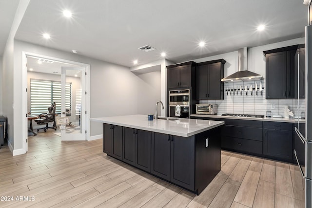 kitchen featuring sink, wall chimney range hood, an island with sink, decorative backsplash, and stainless steel appliances