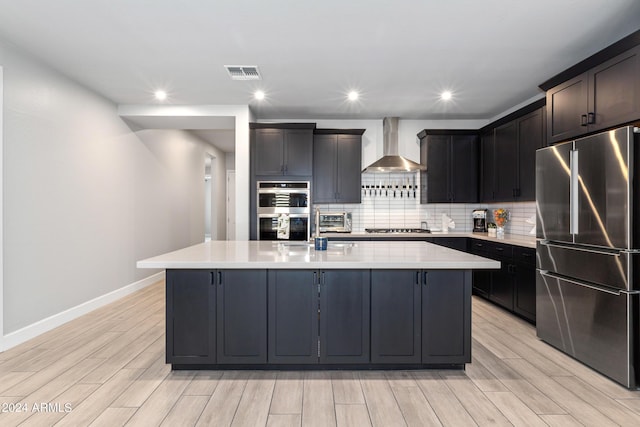 kitchen featuring wall chimney exhaust hood, a center island with sink, decorative backsplash, and appliances with stainless steel finishes