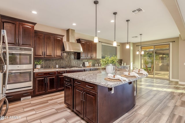 kitchen with a kitchen island, pendant lighting, appliances with stainless steel finishes, tasteful backsplash, and wall chimney range hood