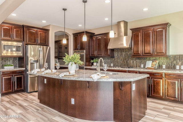 kitchen featuring decorative light fixtures, wall chimney range hood, appliances with stainless steel finishes, and a kitchen island with sink