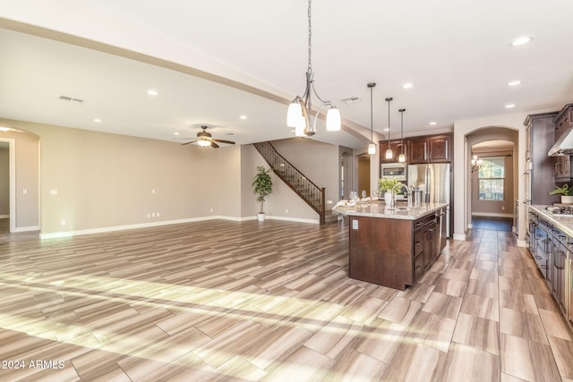 kitchen with light wood-type flooring, light stone counters, ceiling fan with notable chandelier, a kitchen island with sink, and hanging light fixtures