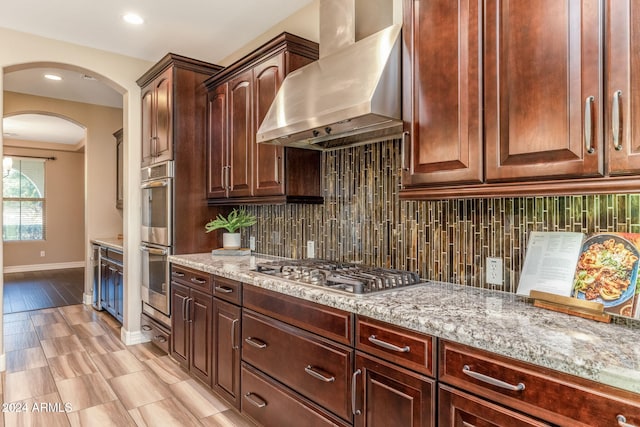 kitchen featuring light stone countertops, backsplash, stainless steel appliances, wall chimney range hood, and light hardwood / wood-style flooring