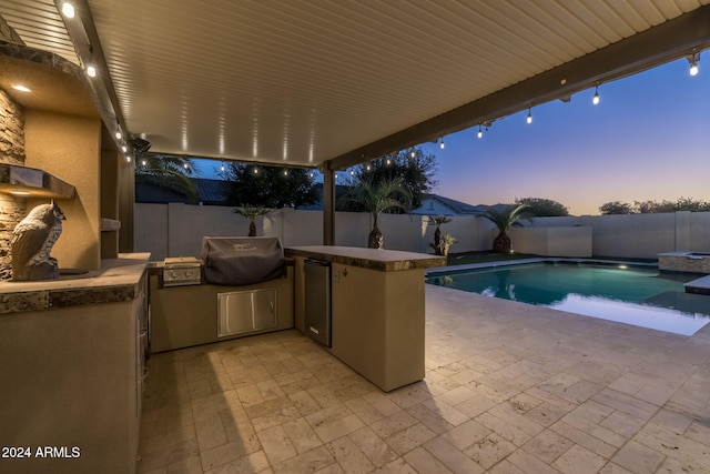 pool at dusk featuring a patio, an outdoor kitchen, and area for grilling