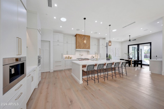 kitchen with a large island with sink, stainless steel oven, hanging light fixtures, white cabinetry, and light hardwood / wood-style floors