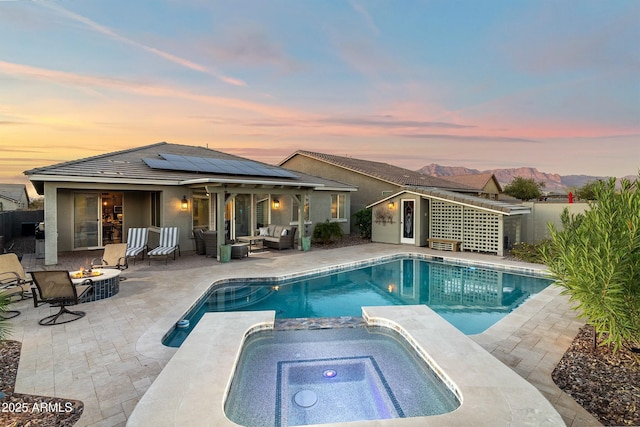 pool at dusk featuring an outdoor living space with a fire pit, a pool with connected hot tub, a patio area, a mountain view, and fence