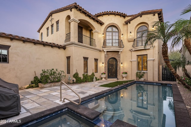 back house at dusk featuring a swimming pool with hot tub, a balcony, and a patio