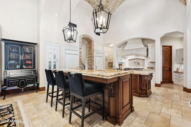 kitchen with hanging light fixtures, a kitchen island with sink, light stone counters, a towering ceiling, and custom exhaust hood