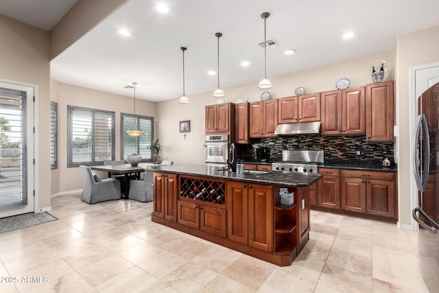 kitchen with pendant lighting, appliances with stainless steel finishes, a kitchen island with sink, dark stone countertops, and tasteful backsplash