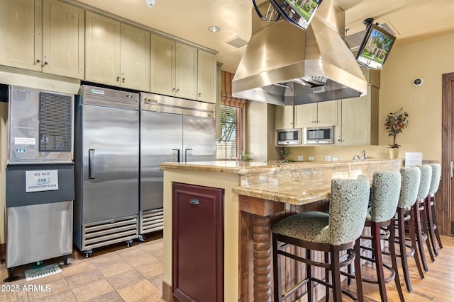 kitchen with built in appliances, island range hood, light stone counters, and cream cabinetry