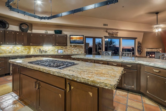 kitchen with light stone counters, dark brown cabinets, a kitchen island, stainless steel gas stovetop, and decorative backsplash