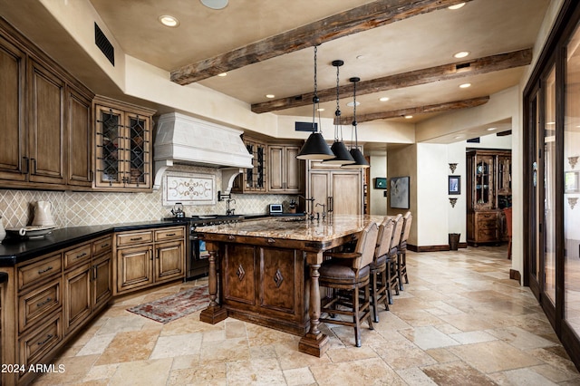 kitchen featuring beam ceiling, backsplash, premium range hood, a kitchen island with sink, and dark stone counters