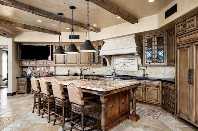 kitchen featuring a sink, stone tile floors, beamed ceiling, custom range hood, and an island with sink