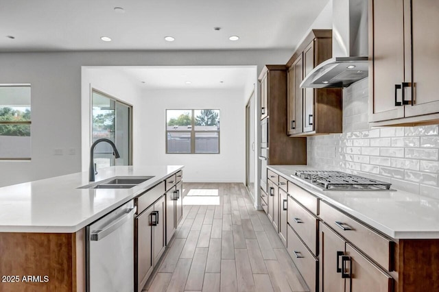 kitchen with light wood-style flooring, a sink, stainless steel appliances, wall chimney exhaust hood, and tasteful backsplash