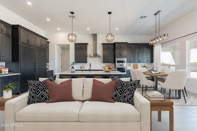 kitchen featuring sink, stainless steel double oven, wall chimney range hood, decorative light fixtures, and a kitchen island with sink