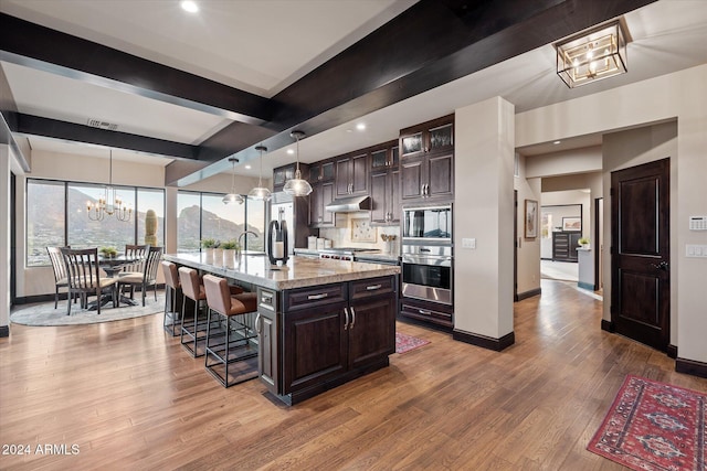 kitchen featuring light stone countertops, appliances with stainless steel finishes, dark brown cabinets, a center island with sink, and hanging light fixtures