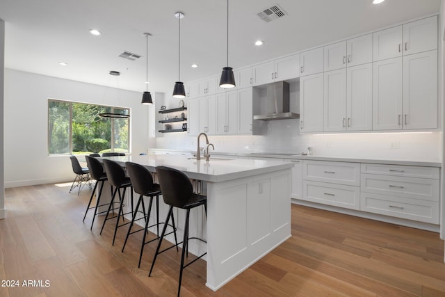 kitchen with light wood-type flooring, white cabinetry, wall chimney exhaust hood, pendant lighting, and a kitchen island with sink