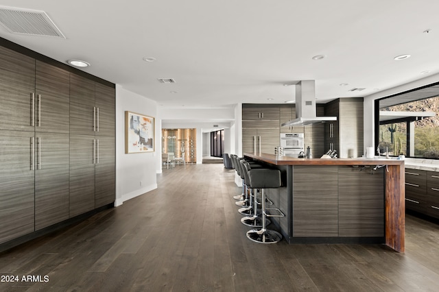 kitchen with wall oven, wall chimney exhaust hood, dark hardwood / wood-style flooring, and a kitchen breakfast bar