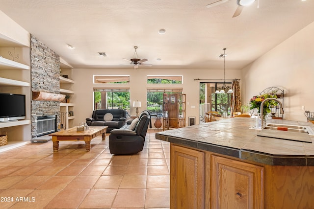 tiled living room featuring built in shelves, ceiling fan, sink, and a stone fireplace